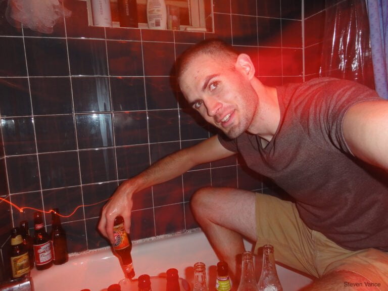 A man sitting in a bathtub surrounded by numerous bottles, holding one bottle and smiling at the camera with red ambient lighting.