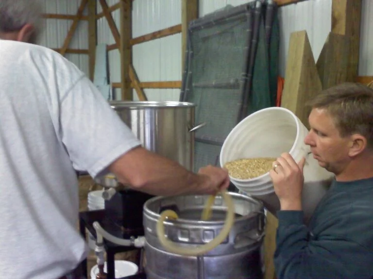 Two men engaged in home brewing, with one pouring grains into a brewing kettle as the other observes.