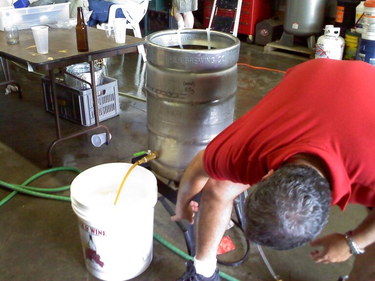 A person in a red shirt bending over to adjust equipment near metal beer kegs in a brewery setting, with hoses, empty containers, and miscellaneous items scattered around.