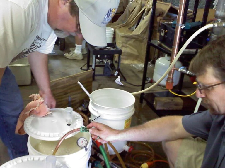 Two men working on a homebrew setup in a garage, with one holding a plastic tube and the other adjusting equipment with gauges and buckets around them.