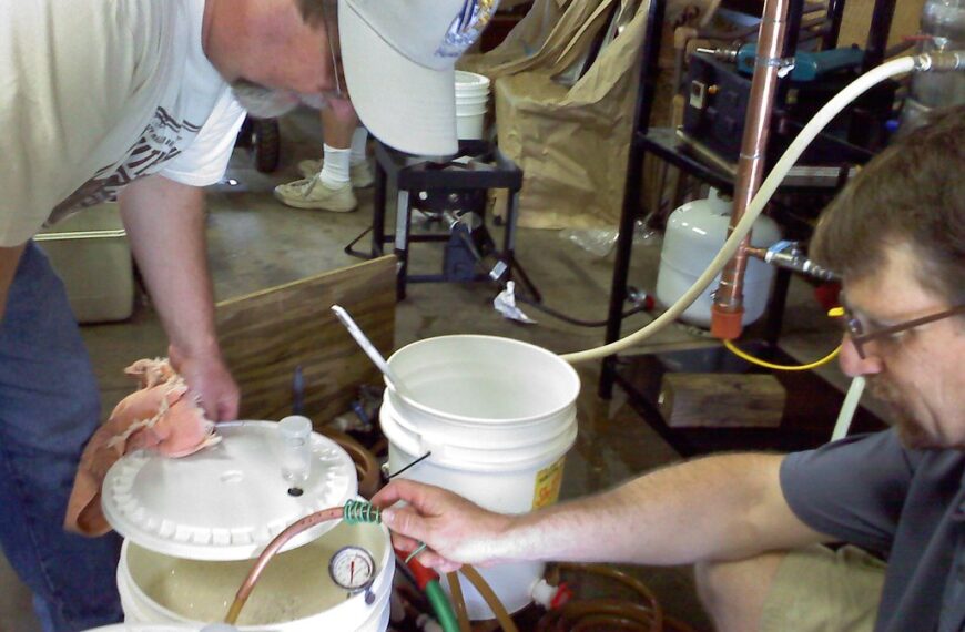 Two men working on a homebrew setup in a garage, with one holding a plastic tube and the other adjusting equipment with gauges and buckets around them.