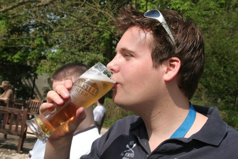 A young man with sunglasses on his head drinking from a glass of beer outdoors.