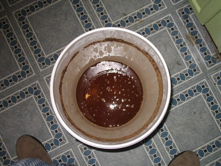 A top-down view of a white bucket filled with dark brown liquid, placed on a tiled floor with a person's feet visible at the bottom edge of the frame.