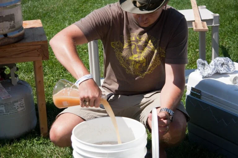 A person in a brown t-shirt and cap is sitting on a chair outdoors, pouring a cloudy liquid from a carboy into a white plastic bucket, with home-brewing equipment nearby.