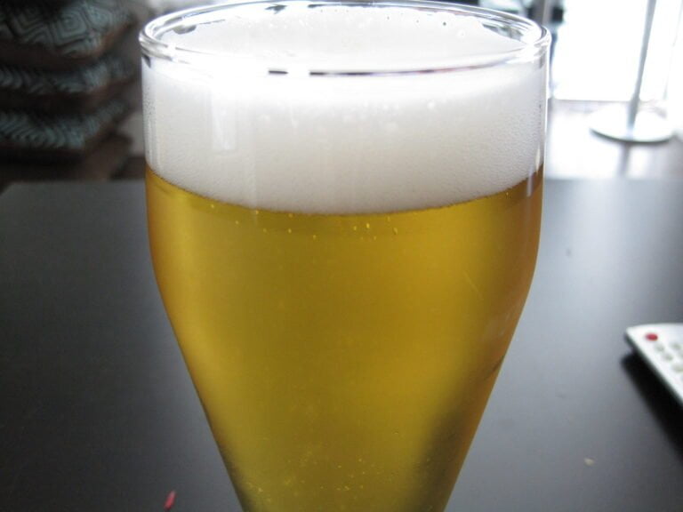 A close-up of a full glass of beer with a frothy head on a table, with vague indoor background details.