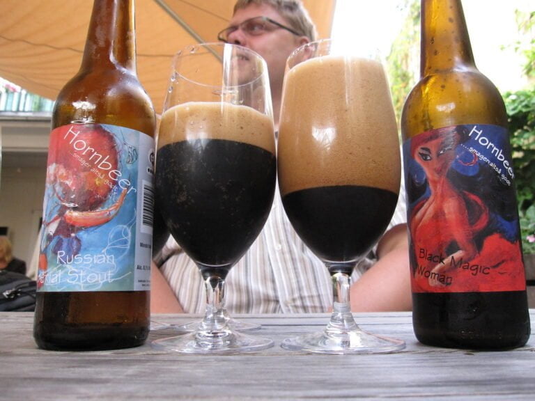 Two dark beers with thick foam are in tulip glasses between their respective beer bottles labeled "Hornbeer Russian Imperial Stout" and "Hornbeer Black Magic Woman" on a table with a person in the background.