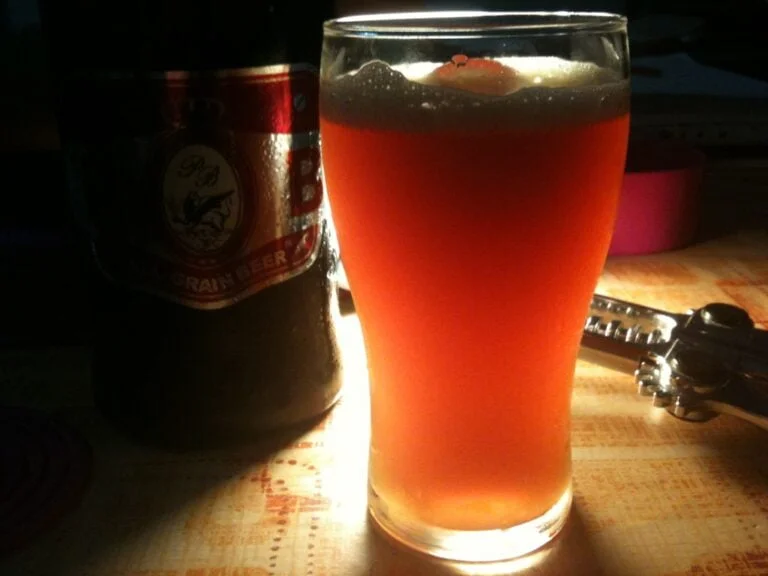 A glass of amber beer backlit by warm light with a beer bottle partially visible on the left, set on a table with indistinct background objects.