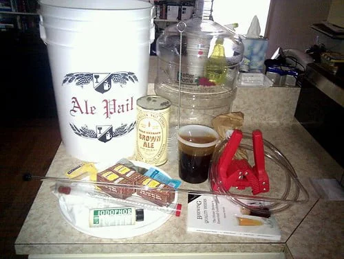 A homebrewing setup with various items including a white fermenting bucket labeled "Ale Pail," a glass carboy, a can of malt extract, packets of brewing yeast, a hydrometer, a siphon tube, a red bottle capper, bottles of additives, and a glass of dark beer.