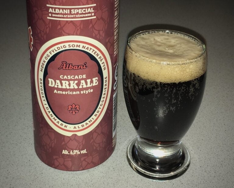 A can of Albani Cascade Dark Ale next to a glass of dark beer with a foamy head on a table.