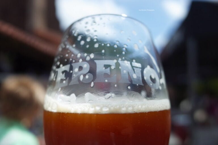 Close-up of a condensation-covered beer glass with the word "BEER" partially visible, filled with amber liquid, set against a blurred background of an outdoor setting.