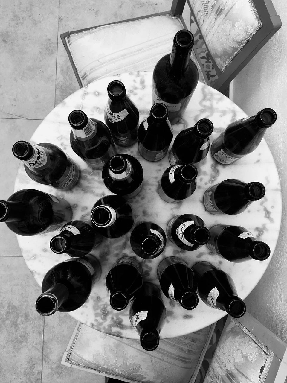 A black and white photograph of multiple empty beer bottles arranged on a round marble table, with a chair partially visible in the background.