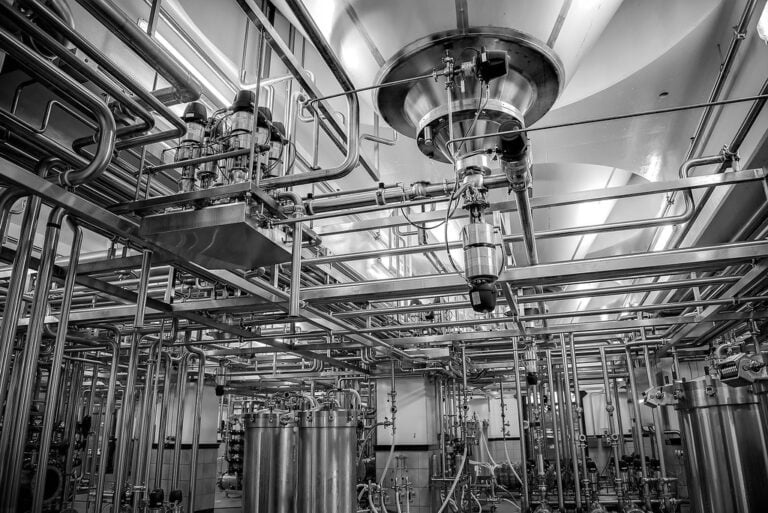 Interior of an industrial facility with a complex arrangement of stainless steel pipes, tanks, and machinery, predominantly in metallic tones.