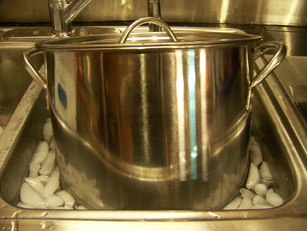 A stainless steel cooking pot with a lid on, sitting in a kitchen sink filled with water and ice cubes for rapid cooling.