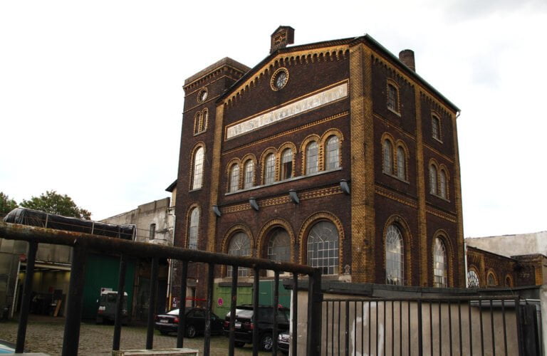 Brick industrial building with ornate facade, arched windows, and a sign near the roof, behind a metal fence with vehicles parked in the foreground under a cloudy sky.