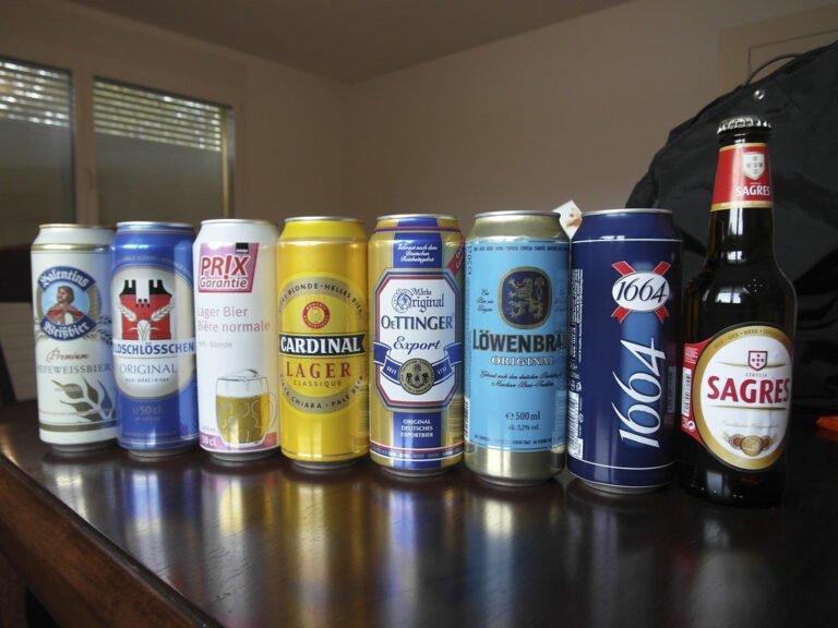 A variety of beer cans and one bottle, with different brands and colors, displayed on a wooden surface inside a room with a window in the background.