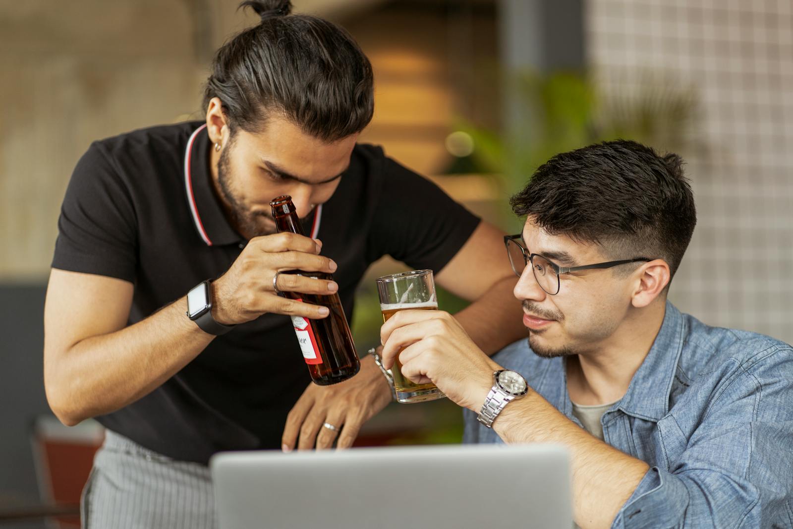 Two men, one drinking from a bottle and the other from a glass, are looking at a laptop screen together in a casual setting.