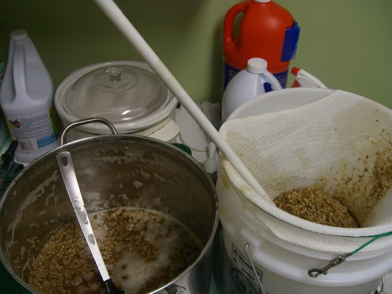 A home brewing setup showing a large pot with grains and liquid on the left, and a fermentation bucket with a cheesecloth bag containing spent grain on the right, amidst various cleaning supplies.