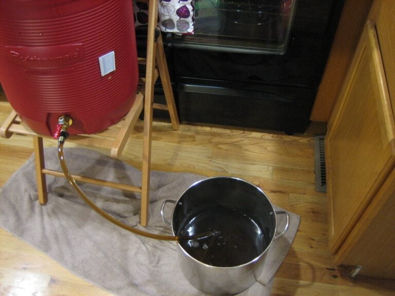 A red water cooler with a spigot sitting on a wooden stand leaking fluid through a hose into a metal pot placed on the floor, with a towel underneath to absorb spills.
