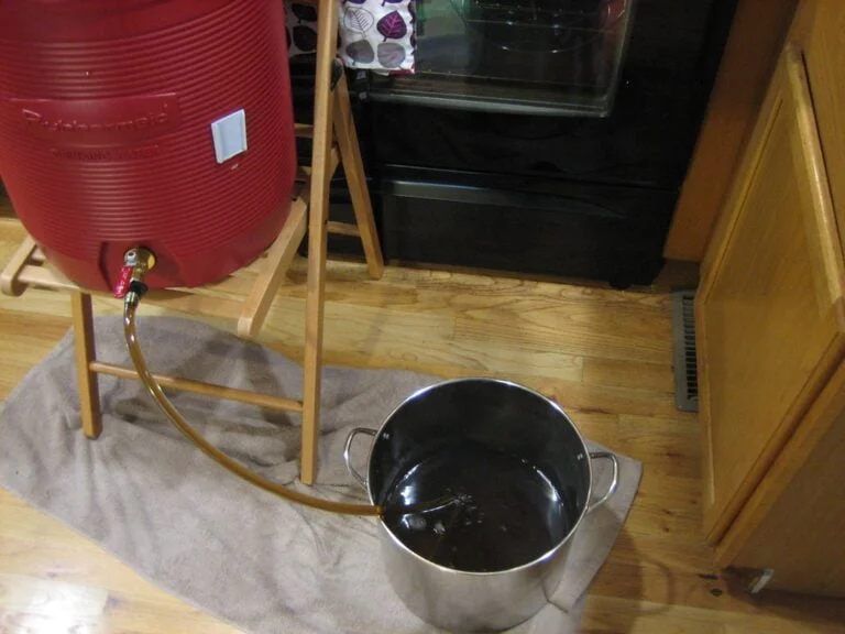 A red water cooler with a spigot sitting on a wooden stand leaking fluid through a hose into a metal pot placed on the floor, with a towel underneath to absorb spills.