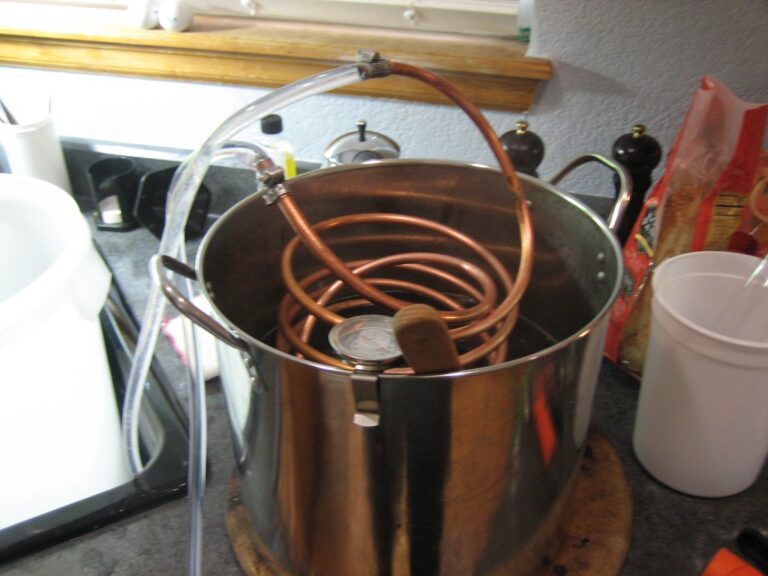 A home distillation setup with a copper coil inside a large stainless steel pot, placed on a kitchen counter among various kitchen utensils and containers.