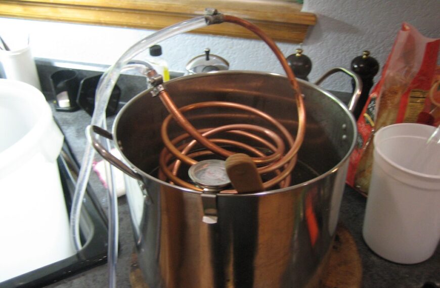 A home distillation setup with a copper coil inside a large stainless steel pot, placed on a kitchen counter among various kitchen utensils and containers.