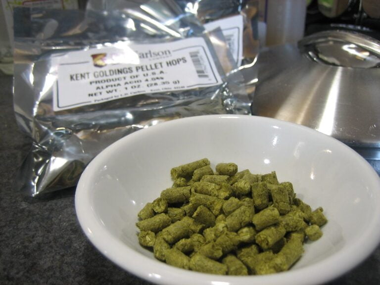 A close-up image of a white bowl filled with pellet hops on a kitchen counter with a blurred package in the background labeled "Kent Goldings Pellet Hops."