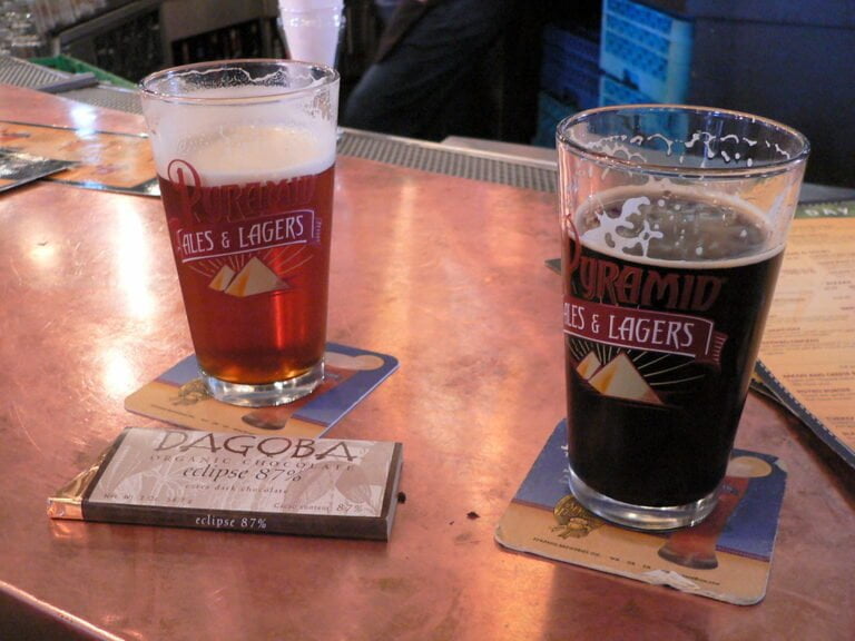 Two glasses of beer on a bar counter, one filled with amber ale and the other with stout, both branded "Pyramid Ales & Lagers." In front lies a bar of "Dagoba Organic Chocolate," and underneath the glasses are matching beer coasters.