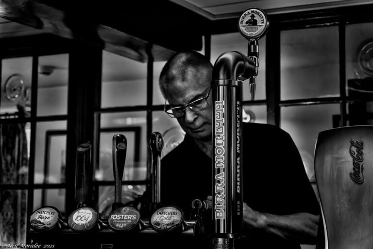A black and white photo of a bartender standing behind a bar with various beer taps in the foreground, including brands like Thatcher's Haze, Foster's, and Birra Moretti, with a Coca-Cola dispenser to the right.