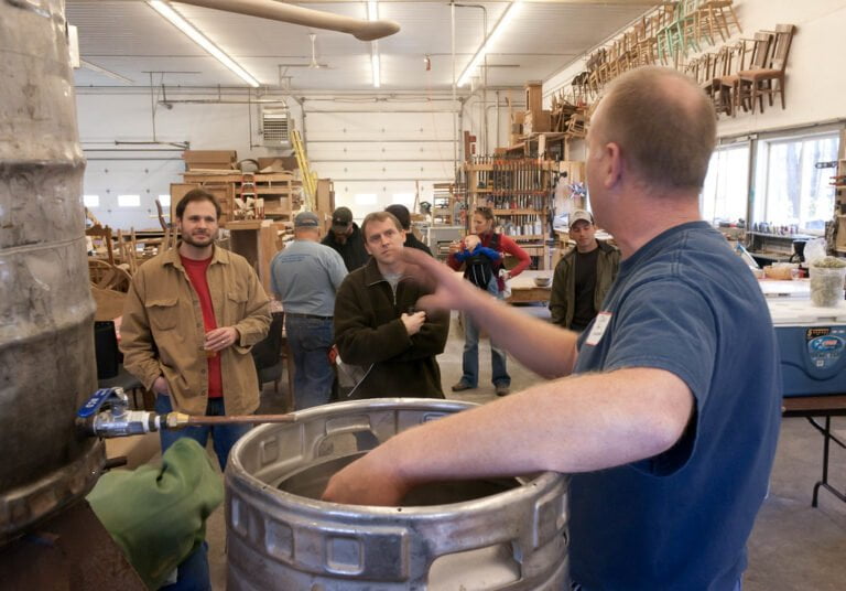 A group of people in a workshop with woodworking materials and chairs hanging on the wall, listening to a man in the foreground who appears to be explaining something, with a metal barrel in the center.