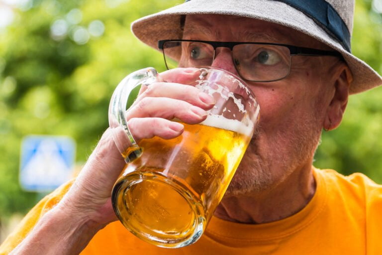 A senior man wearing glasses and a sun hat sipping beer from a large glass mug, with a blurred green background.