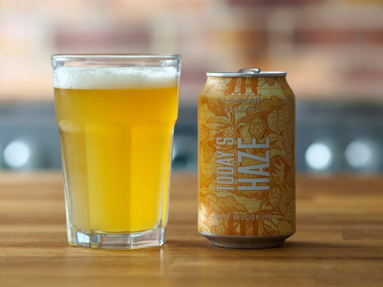 A hazy golden beer in a pint glass next to a Goose Island "Today's Haze" Hazy Session IPA can on a wooden surface.