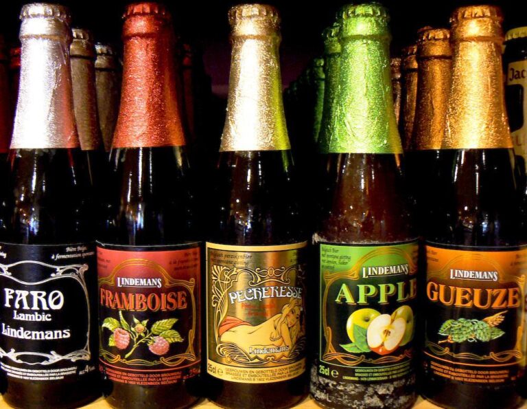 A variety of Lindemans lambic beer bottles with colorful foil caps and labels displaying the flavors: Faro, Framboise, Pêcheresse, Apple, and Gueuze.