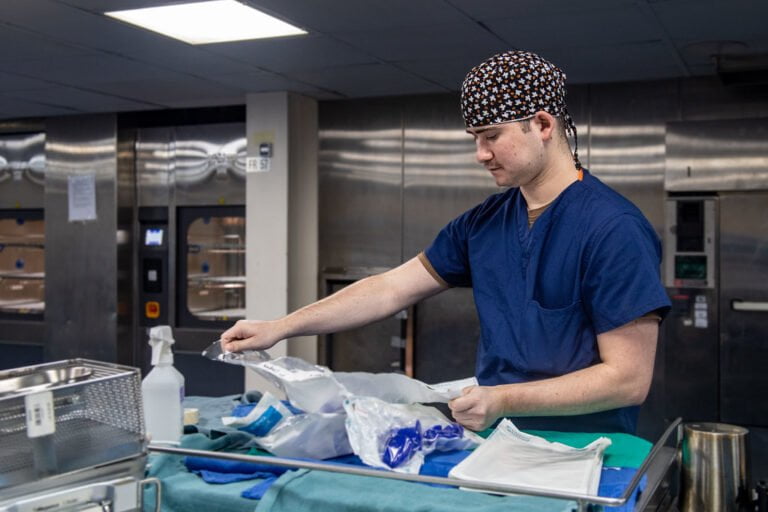 A medical professional in scrubs and a patterned cap carefully arranges sterile instruments on a table in a hospital setting.