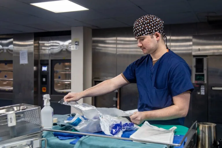 A medical professional in scrubs and a patterned cap carefully arranges sterile instruments on a table in a hospital setting.