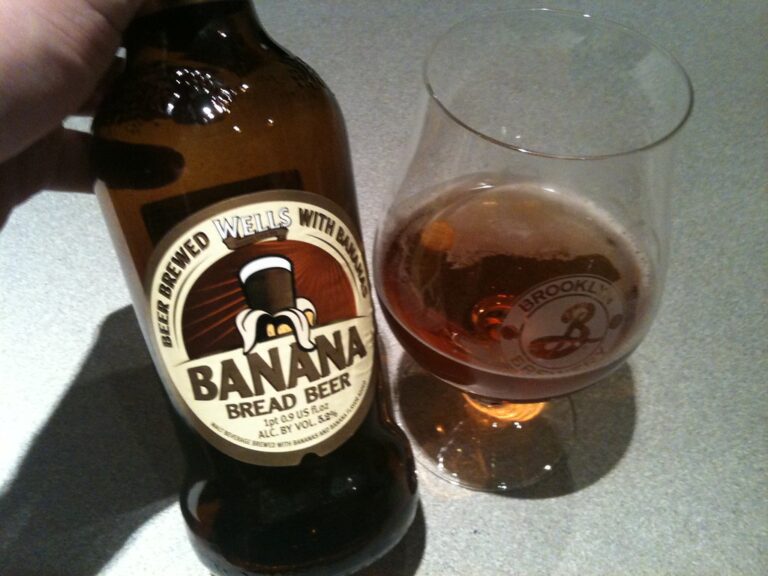 A hand holding a bottle of Wells Banana Bread Beer next to a glass partially filled with beer on a countertop.