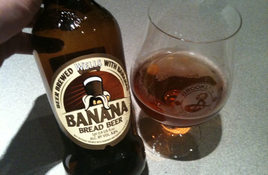 A hand holding a bottle of Wells Banana Bread Beer next to a glass partially filled with beer on a countertop.