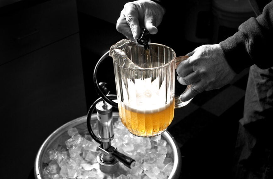 A person pouring beer from a tap into a clear pitcher, with a selective color effect highlighting the golden beer amidst a black and white scene of ice and bar equipment.