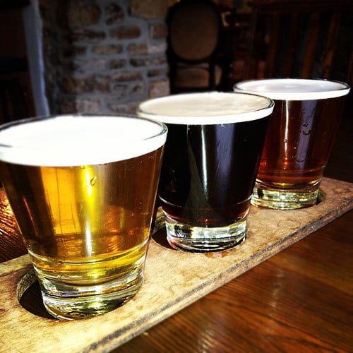 Three glasses of beer with varying colors ranging from light gold to dark brown, served on a wooden paddle board.