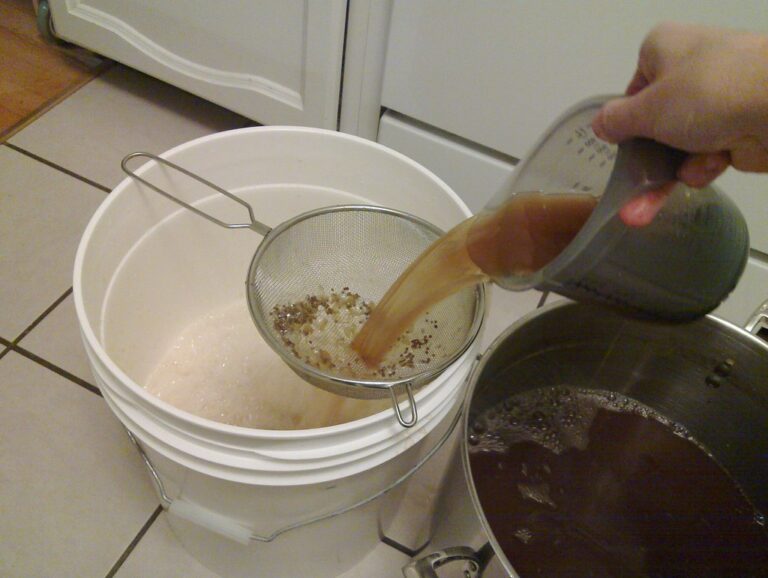A person pouring liquid through a fine mesh strainer into a white bucket; spent grains are visible in the strainer, and a pot with more of the dark liquid is on the right.