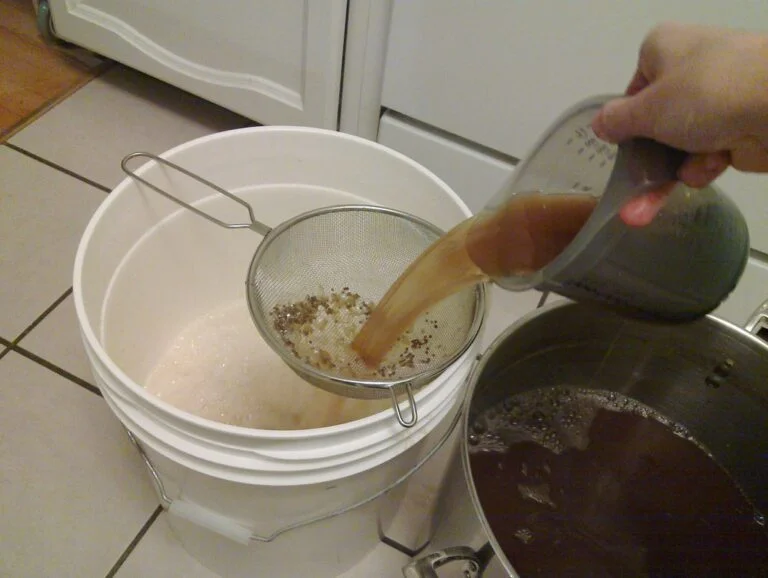 A person pouring liquid through a fine mesh strainer into a white bucket; spent grains are visible in the strainer, and a pot with more of the dark liquid is on the right.