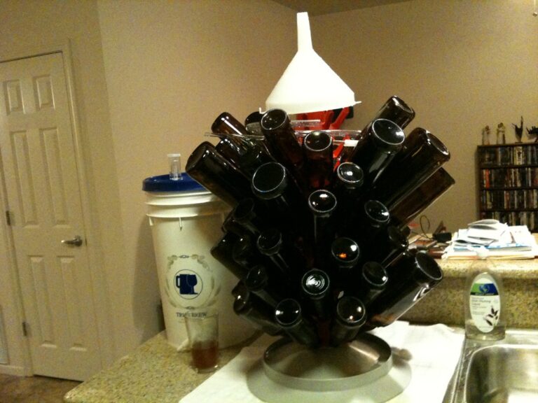 A homebrew drying rack loaded with upside-down brown bottles in a domestic kitchen, with a white bucket and brewing equipment in the background.