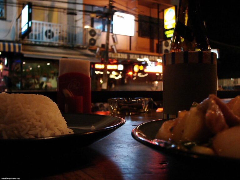 A nighttime street dining scene with a close-up of a plate of white rice, a blurry bottle of beverage with a blank label, and another dish, possibly meat, with a plastic cup and urban background lighting.