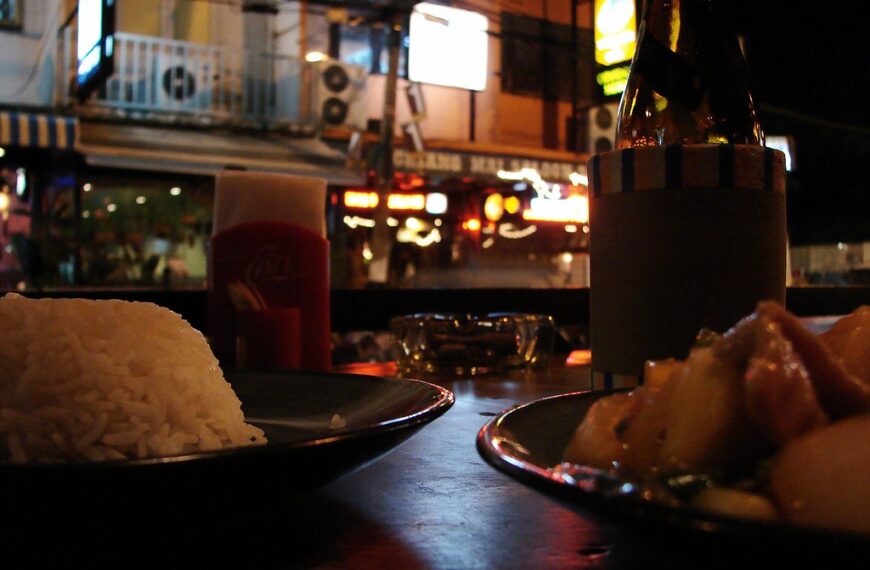 A nighttime street dining scene with a close-up of a plate of white rice, a blurry bottle of beverage with a blank label, and another dish, possibly meat, with a plastic cup and urban background lighting.