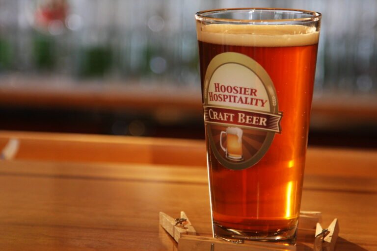 A pint of amber craft beer with a frothy head in a glass with the label "Hoosier Hospitality Craft Beer," sitting on a wooden bar surface, with a blurry background of more glasses.