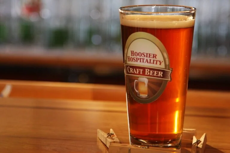 A pint of amber craft beer with a frothy head in a glass with the label "Hoosier Hospitality Craft Beer," sitting on a wooden bar surface, with a blurry background of more glasses.