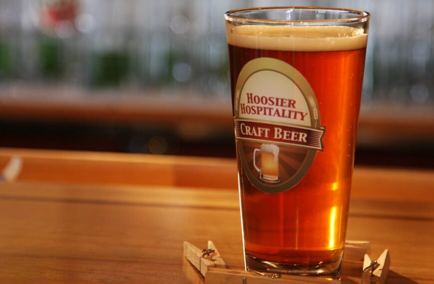 A pint of amber craft beer with a frothy head in a glass with the label "Hoosier Hospitality Craft Beer," sitting on a wooden bar surface, with a blurry background of more glasses.