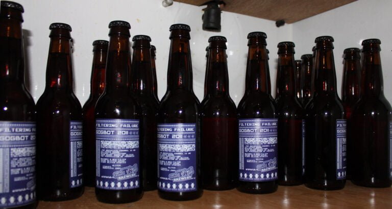 A collection of brown glass beer bottles with blue labels, titled "FILTERING FAILURE," lined up on a white shelf.