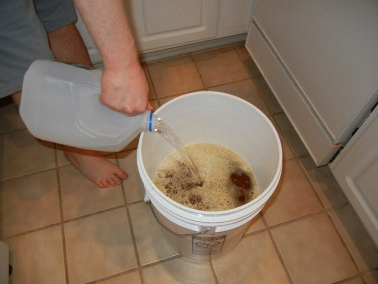 A person is pouring liquid from a gallon jug into a large white bucket filled with foam, standing barefoot on a tiled kitchen floor.