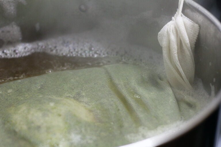 A close-up of a cheesecloth bag being used in the process of brewing or steeping in a pot of simmering liquid with visible steam.