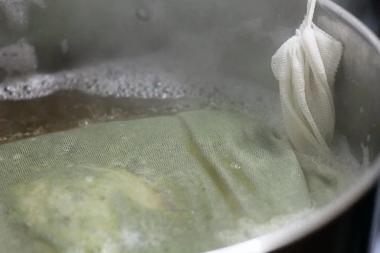 A close-up of a cheesecloth bag being used in the process of brewing or steeping in a pot of simmering liquid with visible steam.